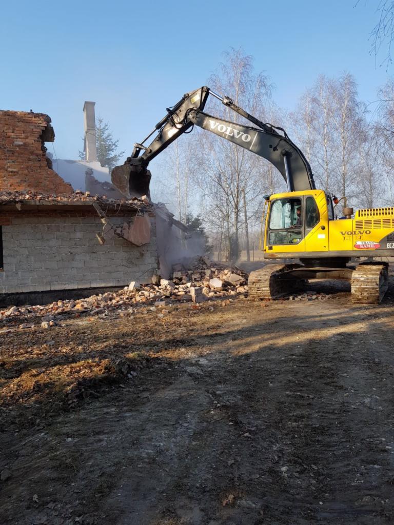 Żółta koparka Volvo na gąsienicach w trakcie rozbiórki murowanego budynku, widoczne gruzowisko, pyląca cegła i fragment komina na tle błękitnego nieba i drzew.