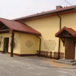 Żółty budynek z czerwonym dachem, zdobiony ludowymi motywami kwiatowymi, z drewnianymi detalami i małym płotkiem.