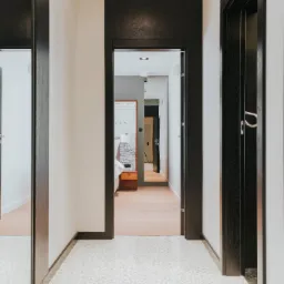 Wnętrze korytarza z ciemnymi, drewnianymi futrynami i lustrem, jasnymi ścianami i mozaikową podłogą, widok w głąb mieszkania.