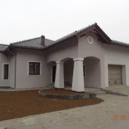 Parterowy dom z beżową elewacją, ciemną dach&oacute;wką i białymi kolumnami portyku, widok z przodu na tle pochmurnego nieba i przygotowanego terenu pod ogr&oacute;d.