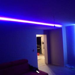 Zakład Remontowo Budowlany Piotr Kogut - Pokój z nowoczesnym oświetleniem LED: listwa sufitowa i pionowa na ścianie, emitujące intensywne niebieskie światło, częściowo widoczny czerwony narożnik sofy w dolnym rogu.