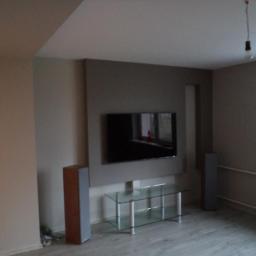 Zakład Remontowo Budowlany Piotr Kogut - Szare wnętrze salonu z telewizorem na ścianie, szklanym stolikiem RTV i dwoma głośnikami; surowa żarówka zwisa z sufitu.