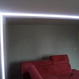 Zakład Remontowo Budowlany Piotr Kogut - Nowoczesny salon z czerwonym narożnikiem i podświetlanym sufitem LED, widok na otwarte drzwi do kuchni.