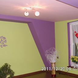 Zakład Remontowo Budowlany Piotr Kogut - Pokój z jasnozielonymi i fioletowymi ścianami, dekoracyjną naklejką drzewa, ceglanym narożnikiem, oknem z figurką Świętego Mikołaja i bukietem żółtych kwiatów.