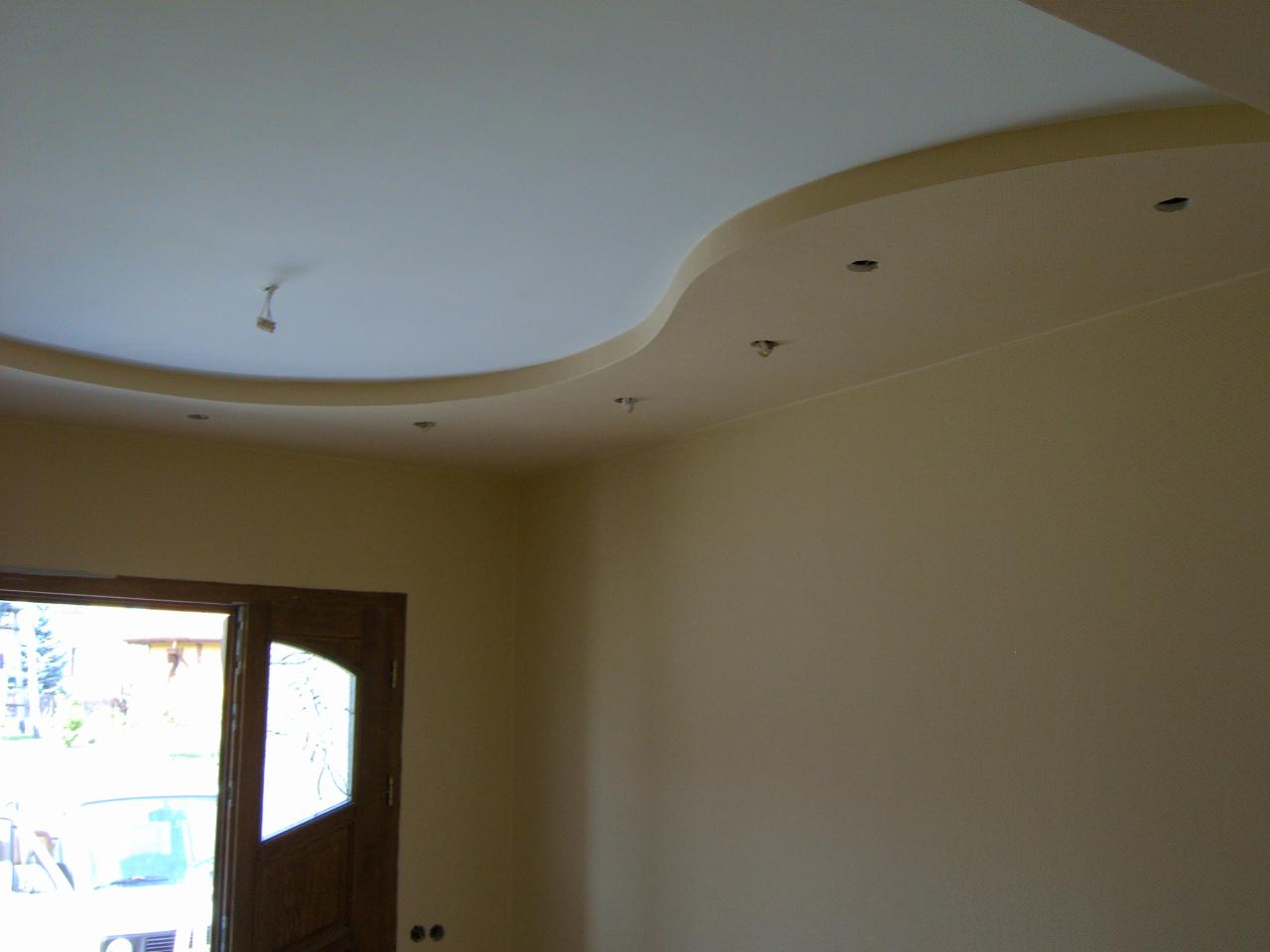 Kremowy sufit podwieszany z falistym obniżeniem i oczkami pod oświetlenie punktowe, widok z narożnika pokoju z otwartymi drzwiami.