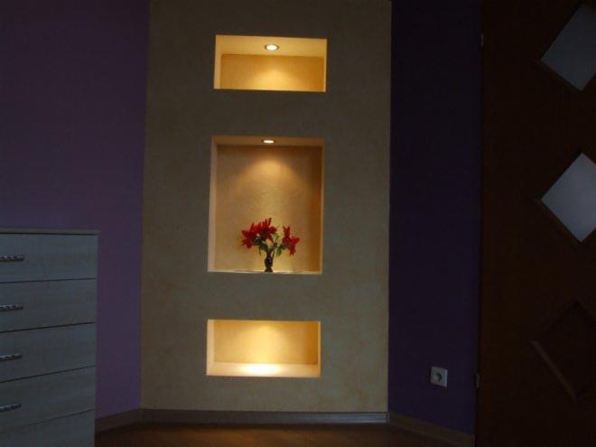 Wnęki ścienne z oświetleniem punktowym, środkowa wnęka z wazonem czerwonych kwiatów na tle fioletowej ściany i komody