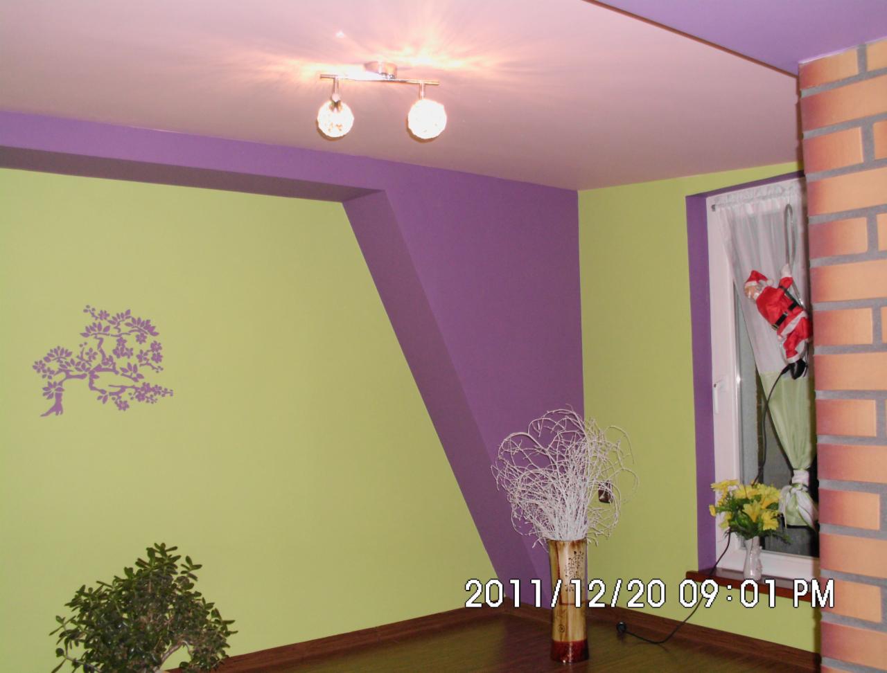 Pokój z jasnozielonymi i fioletowymi ścianami, dekoracyjną naklejką drzewa, ceglanym narożnikiem, oknem z figurką Świętego Mikołaja i bukietem żółtych kwiatów.