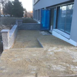 Taras z szarej kostki brukowej w trakcie budowy, zasypany piaskiem, z niskim murem oporowym z betonowych bloczków i wejściem do budynku zabezpieczonym folią.