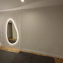 Ściana z dekoracyjnymi panelami pionowymi i podświetlanym lustrem o organicznym kształcie, parkiet w jodełkę na podłodze, widok na drzwi w głębi.