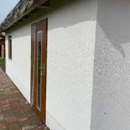 Boczny widok budynku z jasną elewacją o fakturze baranka, brązowe drzwi z szybą zabezpieczone folią, kostka brukowa prowadząca do drewnianej szopy w tle, pochmurne niebo.