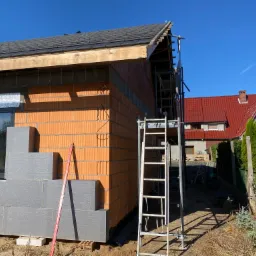 Wznoszenie ścian z pustaków ceramicznych i częściowe ocieplenie fundamentów szarymi płytami izolacyjnymi, widoczna poziomica i rusztowanie na tle błękitnego nieba.