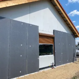 Montaż szarych płyt izolacyjnych na ścianie zewnętrznej domu jednorodzinnego, widoczne białe kołki mocujące, okno z brązową roletą, dach z czerwoną dachówką, piasek na ziemi.