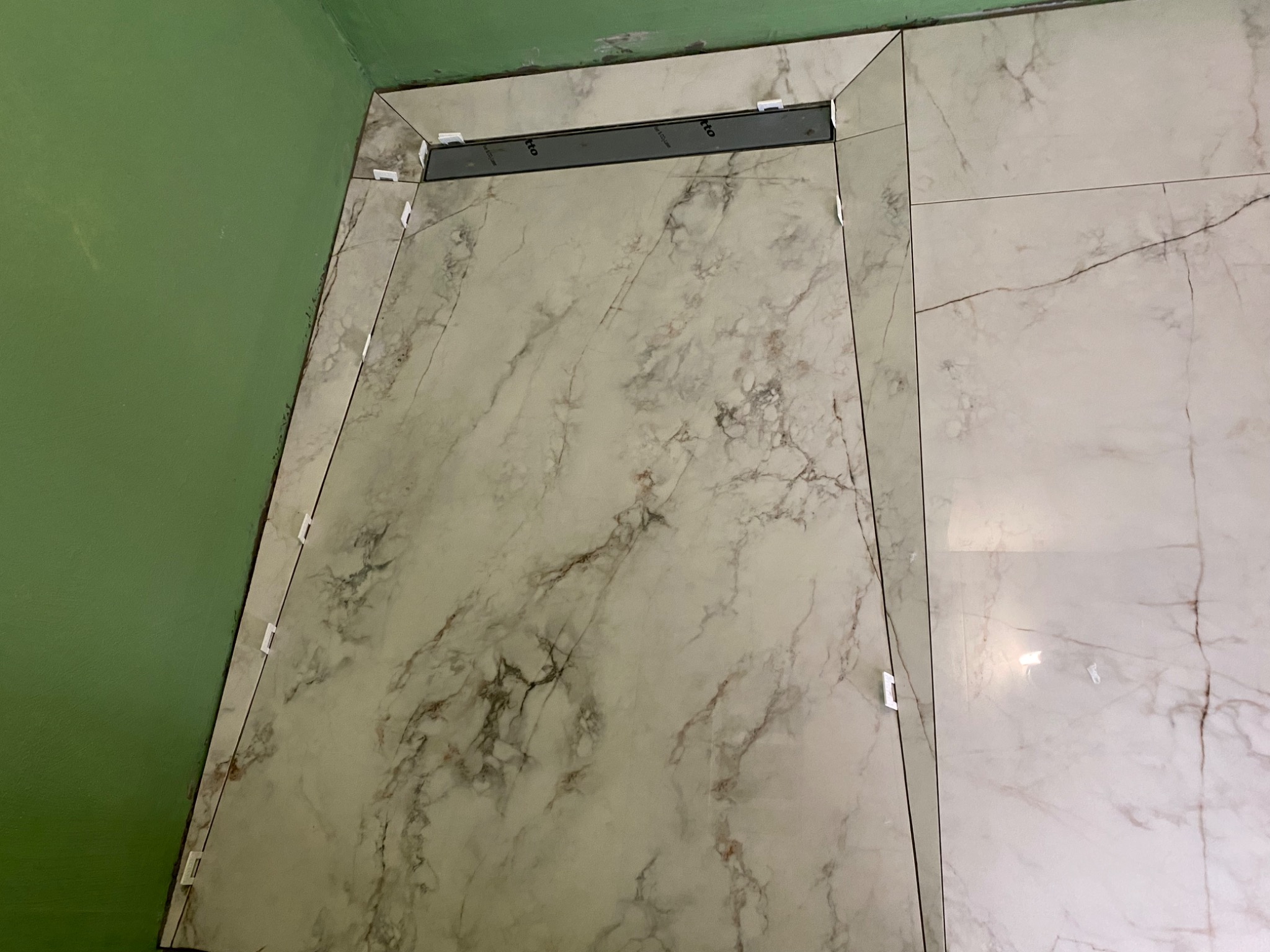 Ułożone płytki podłogowe imitujące marmur w łazience, z widocznym odpływem liniowym i plastikowymi krzyżykami dystansowymi przy ścianie pomalowanej na zielono.