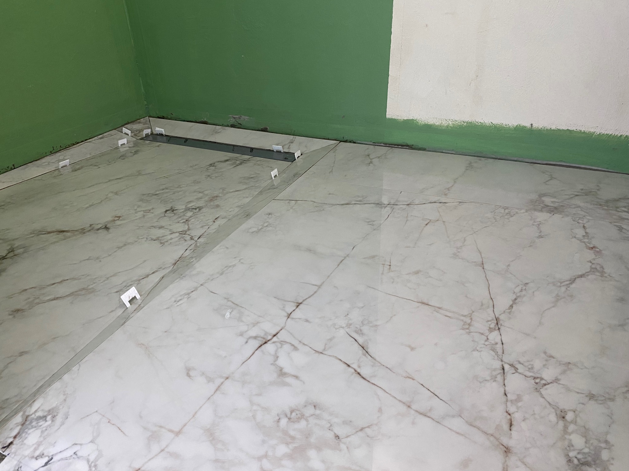 Układanie płytek podłogowych imitujących marmur, z użyciem klipsów poziomujących, w rogu pomieszczenia z zieloną i częściowo pomalowaną ścianą.