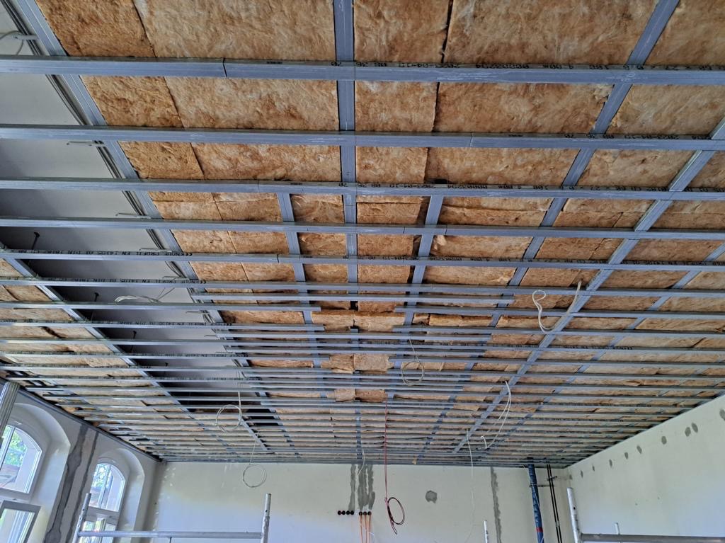 Sufit podwieszany w trakcie montażu z widoczną konstrukcją metalową i izolacją z wełny mineralnej, częściowo zamontowane okablowanie elektryczne, widok z dołu.
