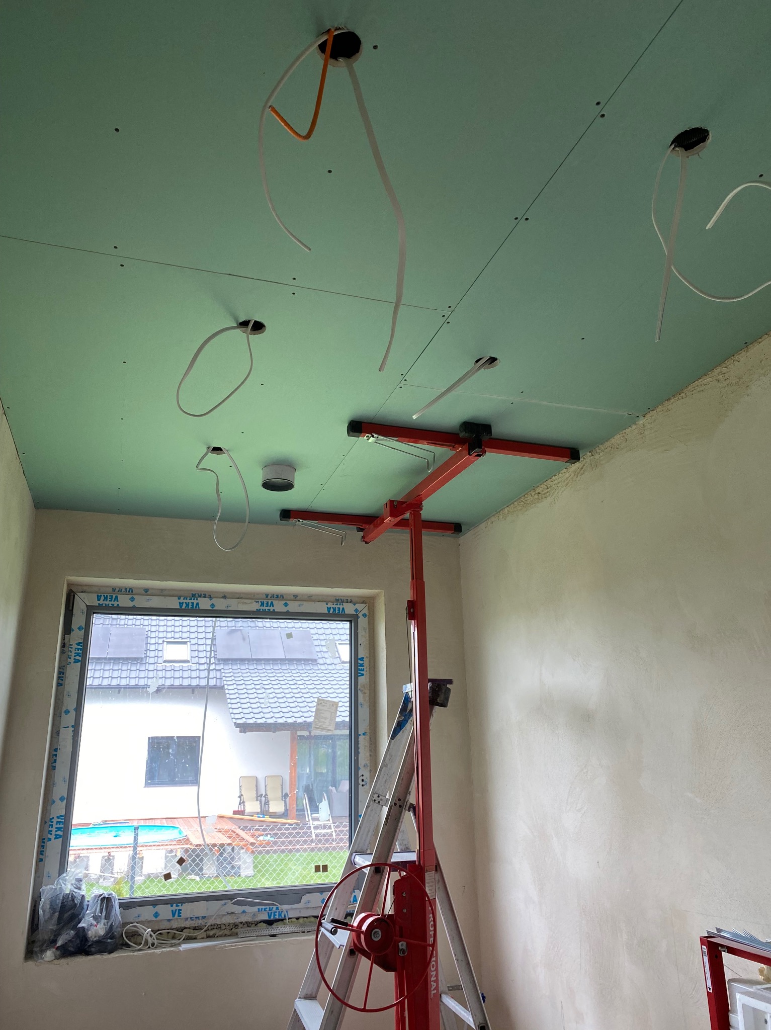 Montaż sufitu podwieszanego: widok na częściowo zamontowane płyty na stelażu, z widocznymi przewodami elektrycznymi wystającymi z otworów, podnośnik do płyt gipsowych, oraz okno z widokiem na dom...