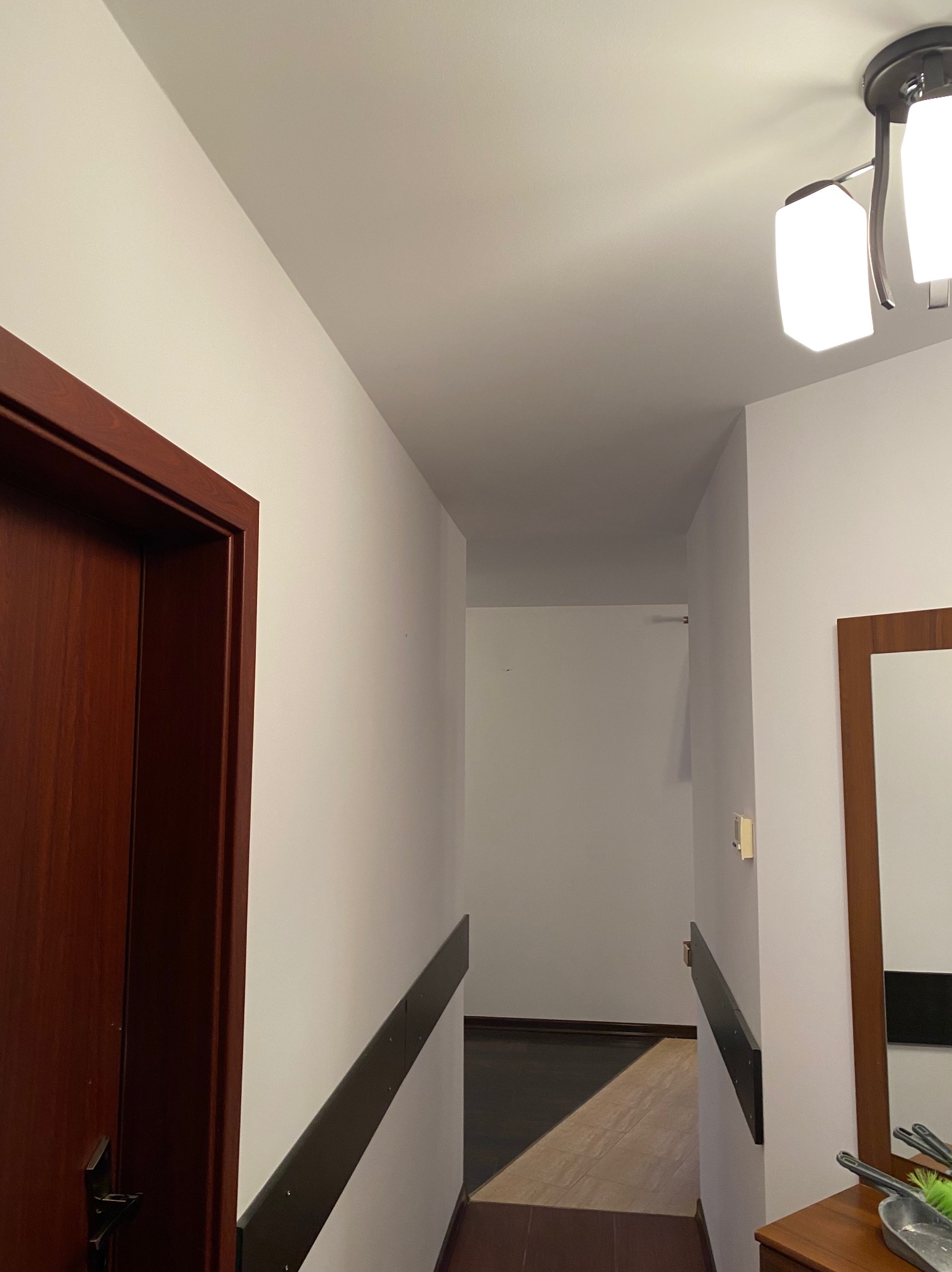 Wnętrze korytarza po remoncie: białe ściany, ciemna listwa przypodłogowa, drewniane drzwi i rama lustra, widoczna część podłogi z dwoma rodzajami paneli, oświetlenie sufitowe.