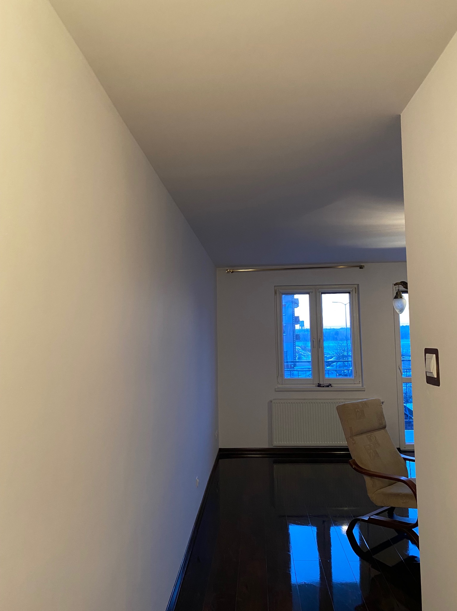 Pomalowane na biało ściany i sufit w pokoju z ciemną podłogą, widok na okno z grzejnikiem i krzesłem w tle, widoczne gniazdko elektryczne na ścianie.