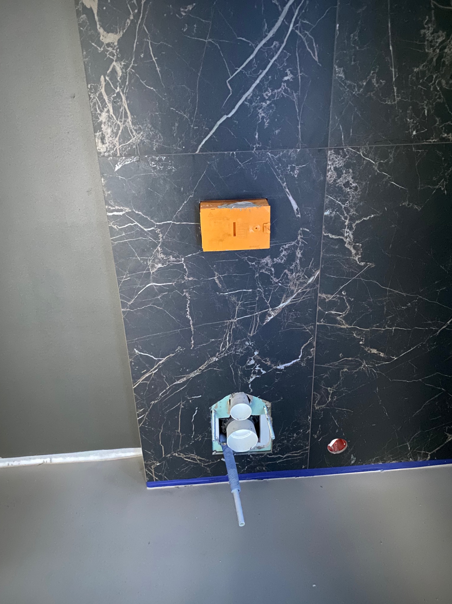 Montaż podtynkowego systemu WC na ścianie z czarnych płytek imitujących marmur, widoczne elementy instalacji i pomarańczowa obudowa stelaża.