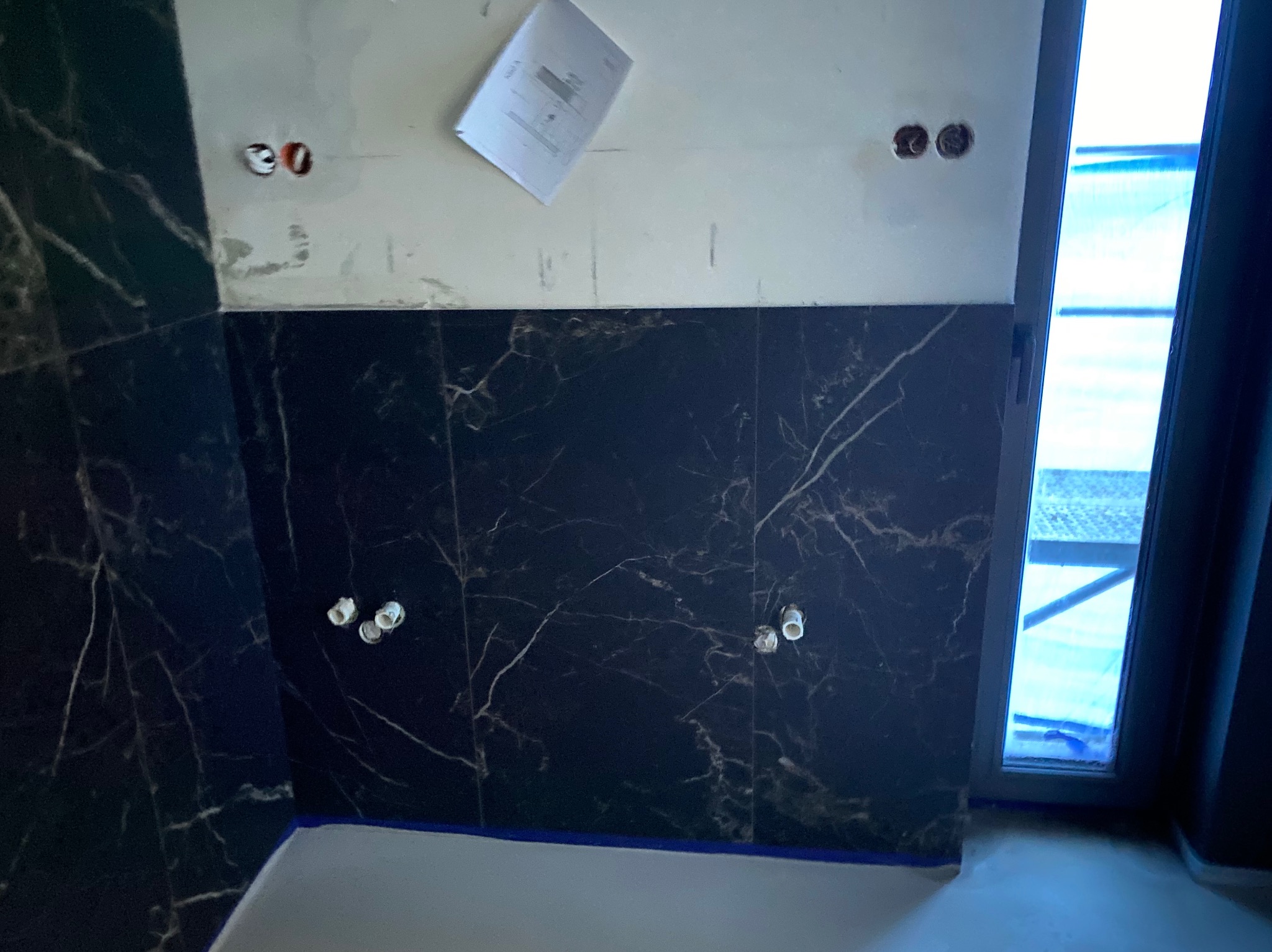 Częściowo wykończona ściana łazienki z ciemnymi płytkami imitującymi marmur, widoczne otwory na instalacje, zabezpieczona taśmą krawędź wanny i dokument przyklejony do górnej części ściany.