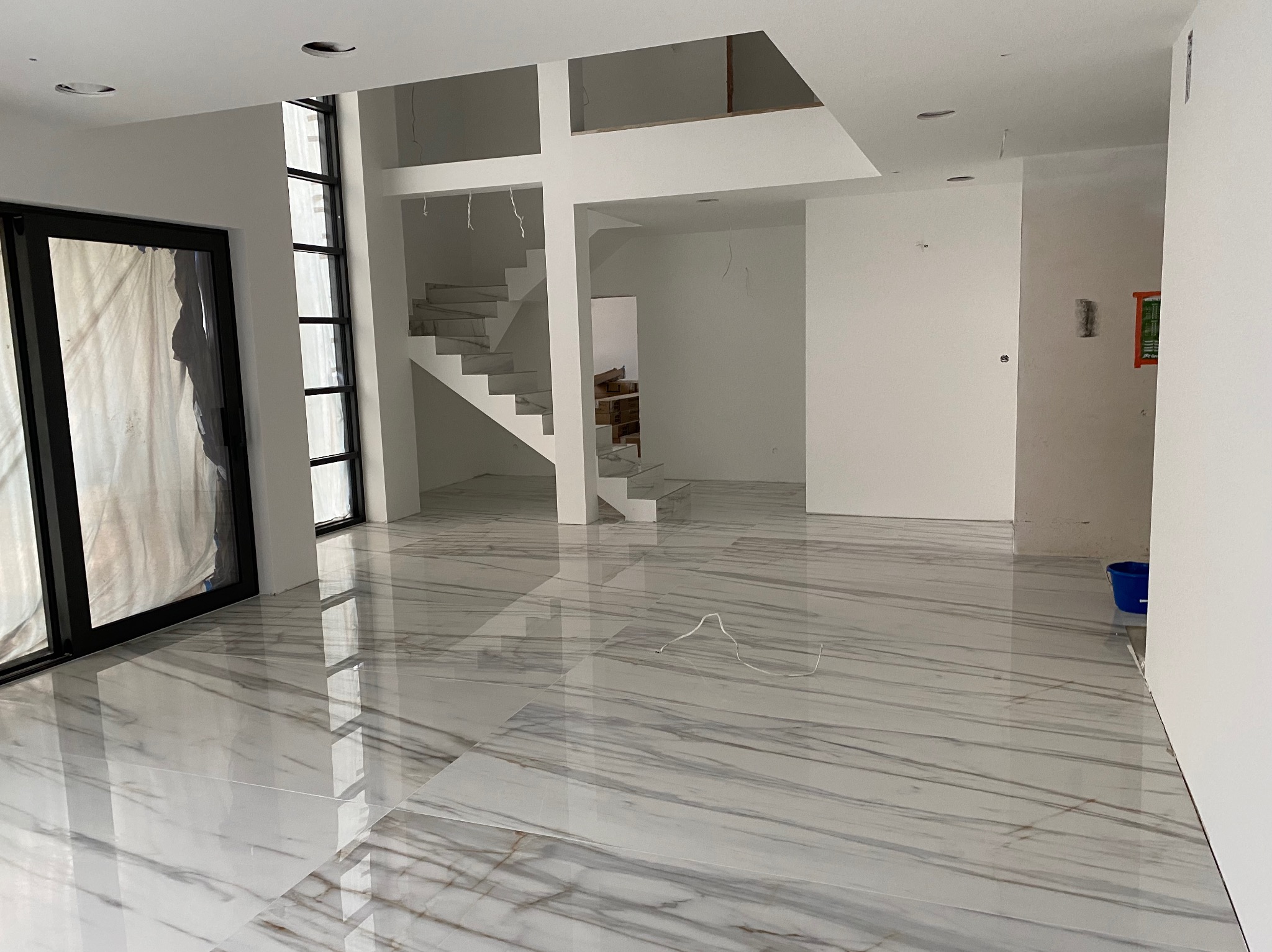 Wnętrze domu w trakcie wykończenia z marmurową podłogą, widoczne schody i okna z czarnymi ramami, zabezpieczone folią. Otwarta przestrzeń z białymi ścianami, przygotowana do dalszych prac.