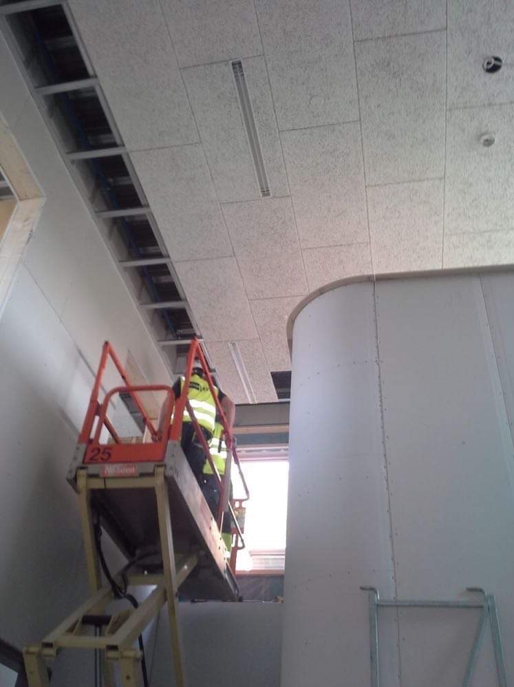 Pracownik w kamizelce odblaskowej na podnośniku montuje sufit podwieszany z płyt o nieregularnej fakturze, widoczne metalowe profile konstrukcyjne i fragment ściany z zaokrąglonym narożnikiem.