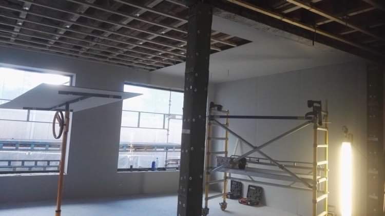 Wnętrze w trakcie prac wykończeniowych: widoczna konstrukcja sufitowa, częściowo zamontowane płyty na ścianach, podnośnik do płyt z zamontowaną płytą, rusztowanie i oświetlenie budowlane.