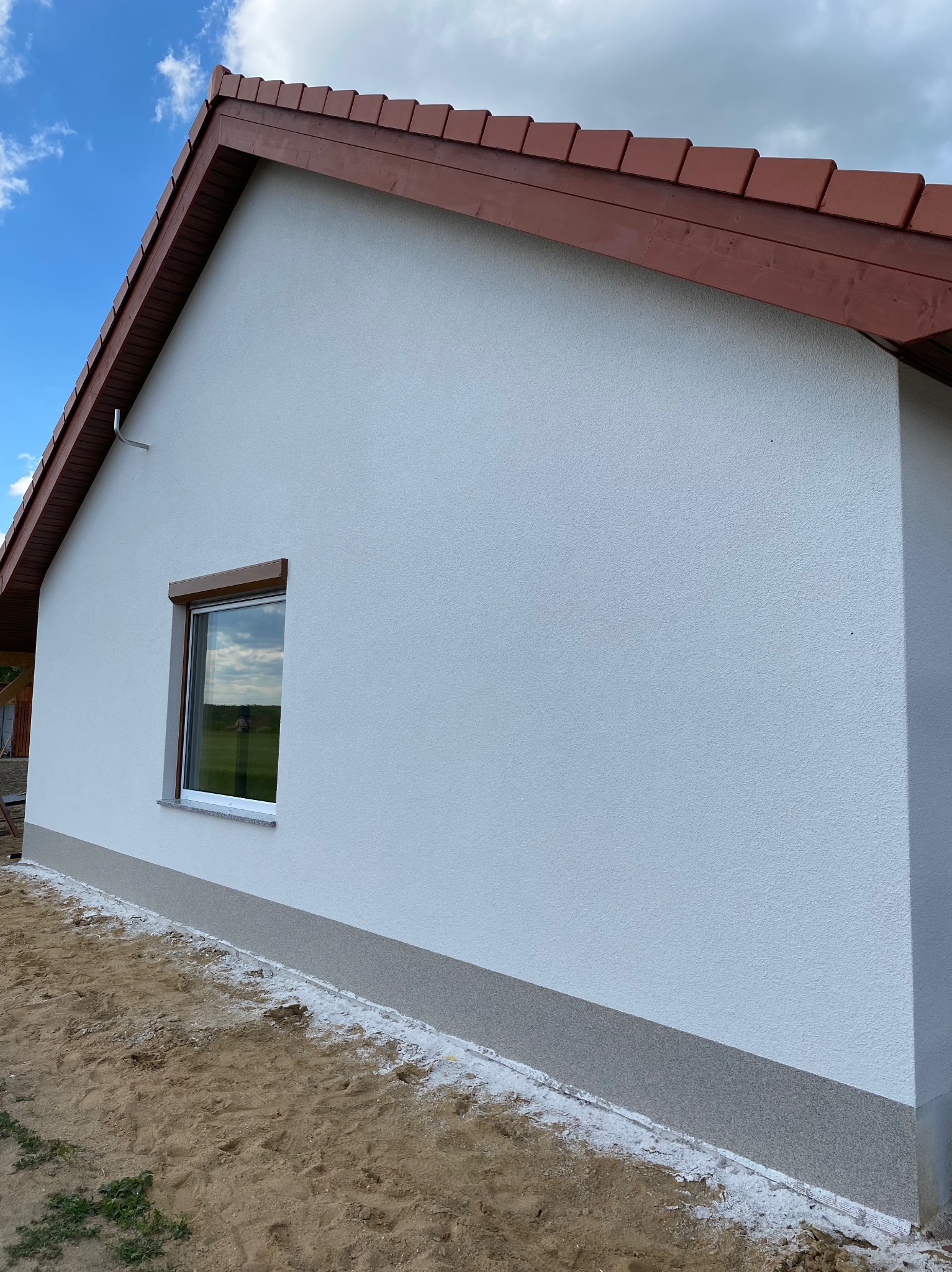 Fragment ściany domu jednorodzinnego w Ścinawie po tynkowaniu elewacji z widocznym oknem z brązowymi roletami i fragmentem dachu z brązową dachówką. Wzdłuż ściany widoczny pas szarego tynku.