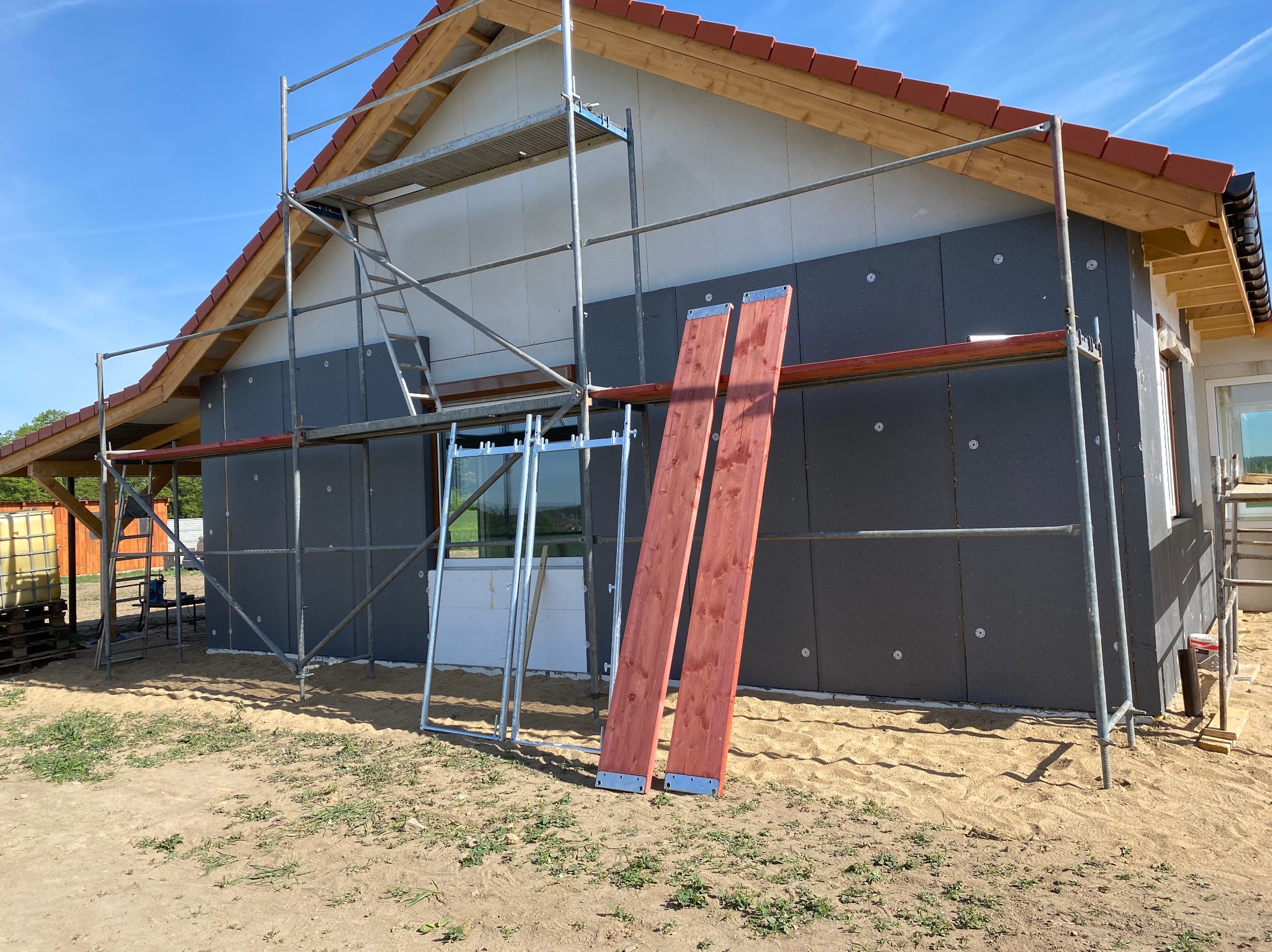 Budowa domu szkieletowego z ociepleniem elewacji styropianem grafitowym, widoczne rusztowanie i dwie deski z metalowymi mocowaniami na ziemi.