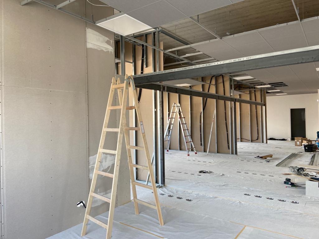 Montaż ścian działowych: widok na metalową konstrukcję i częściowo zamontowane płyty na ścianach, z drewnianą drabiną w pierwszym planie i folią ochronną na podłodze.