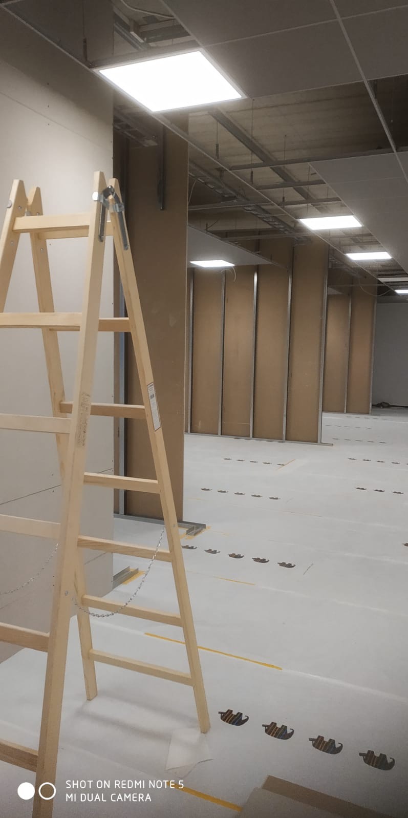 Wnętrze w trakcie montażu ścian działowych z płyt gipsowo-kartonowych, widoczna drewniana drabina oparta o ścianę, metalowe profile konstrukcyjne oraz wzór na podłodze zabezpieczonej folią.