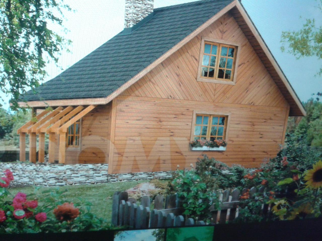 Niewielki, parterowy budynek z bali z ciemną dachówką, ozdobiony kwiatami w skrzynkach okiennych i drewnianą pergolą nad wejściem, otoczony ogrodem z niskim płotkiem.