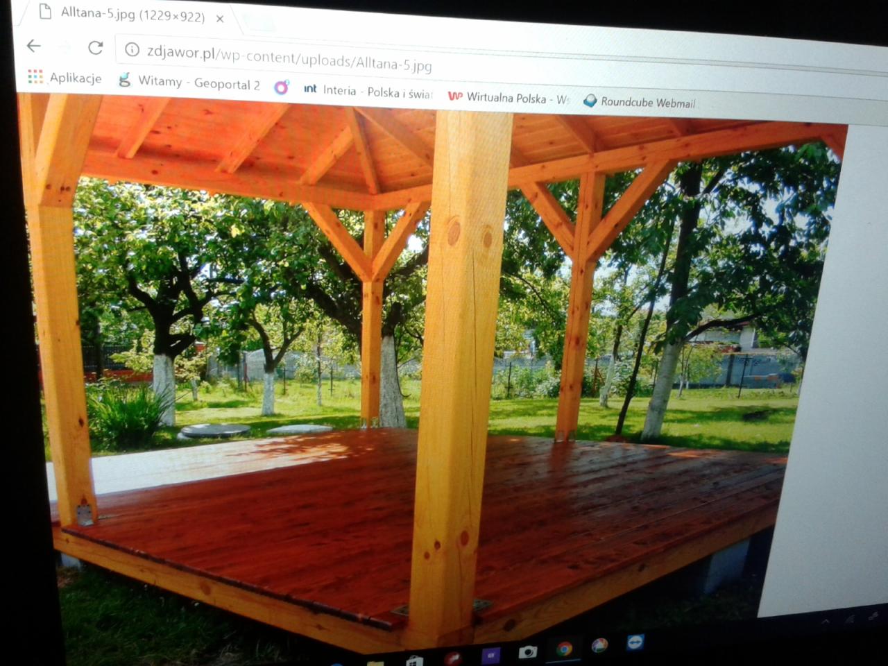 Drewniana altana z bejcowaną podłogą na tle zielonego ogrodu z drzewami o bielonych pniach, widok z zewnątrz.