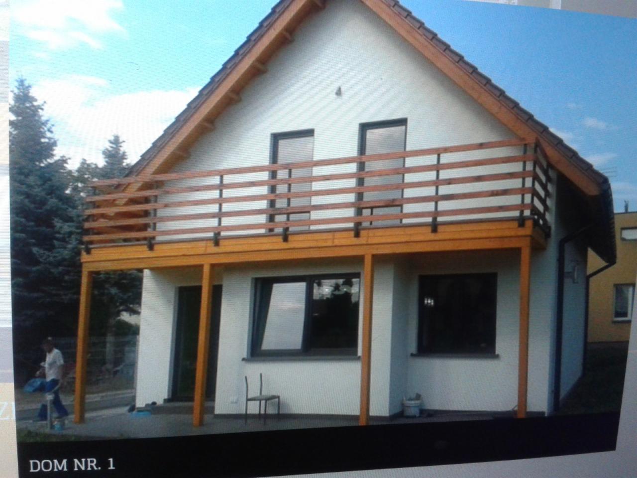 Dwukondygnacyjny dom z białymi ścianami i drewnianymi elementami wykończeniowymi, balkon z drewnianą balustradą na piętrze, widoczna osoba z torbą w tle.
