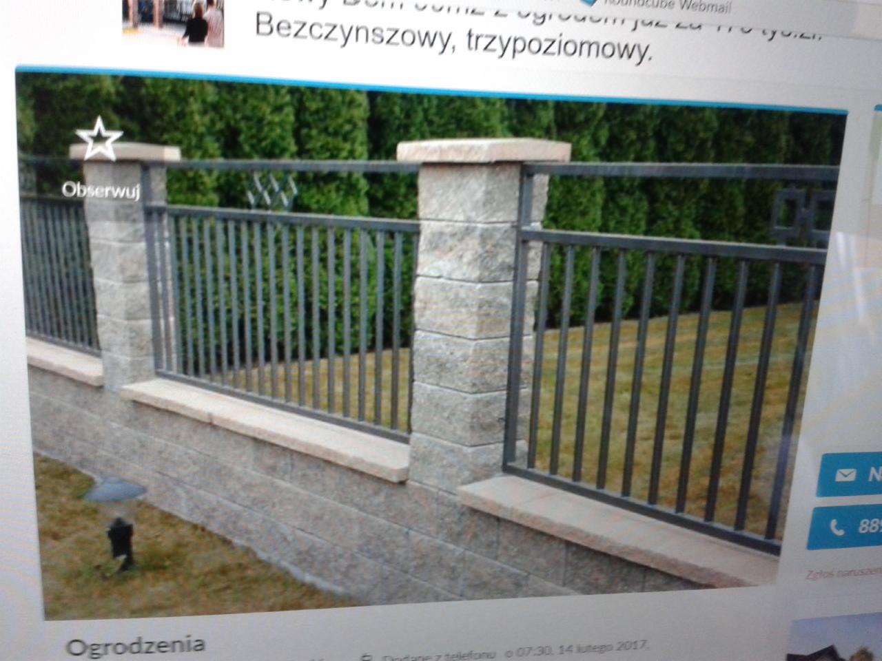 Fragment ogrodzenia z szarej cegły i metalowych prętów, zwieńczonego kamiennymi płytami, z lampą ogrodową w tle i zielonymi tujami w głębi.