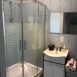 Łazienka z kabiną prysznicową z matowymi poziomymi paskami, umywalką z szafką, lustrem, kwiatem w doniczce, ręcznikami i szarymi płytkami na ścianach.