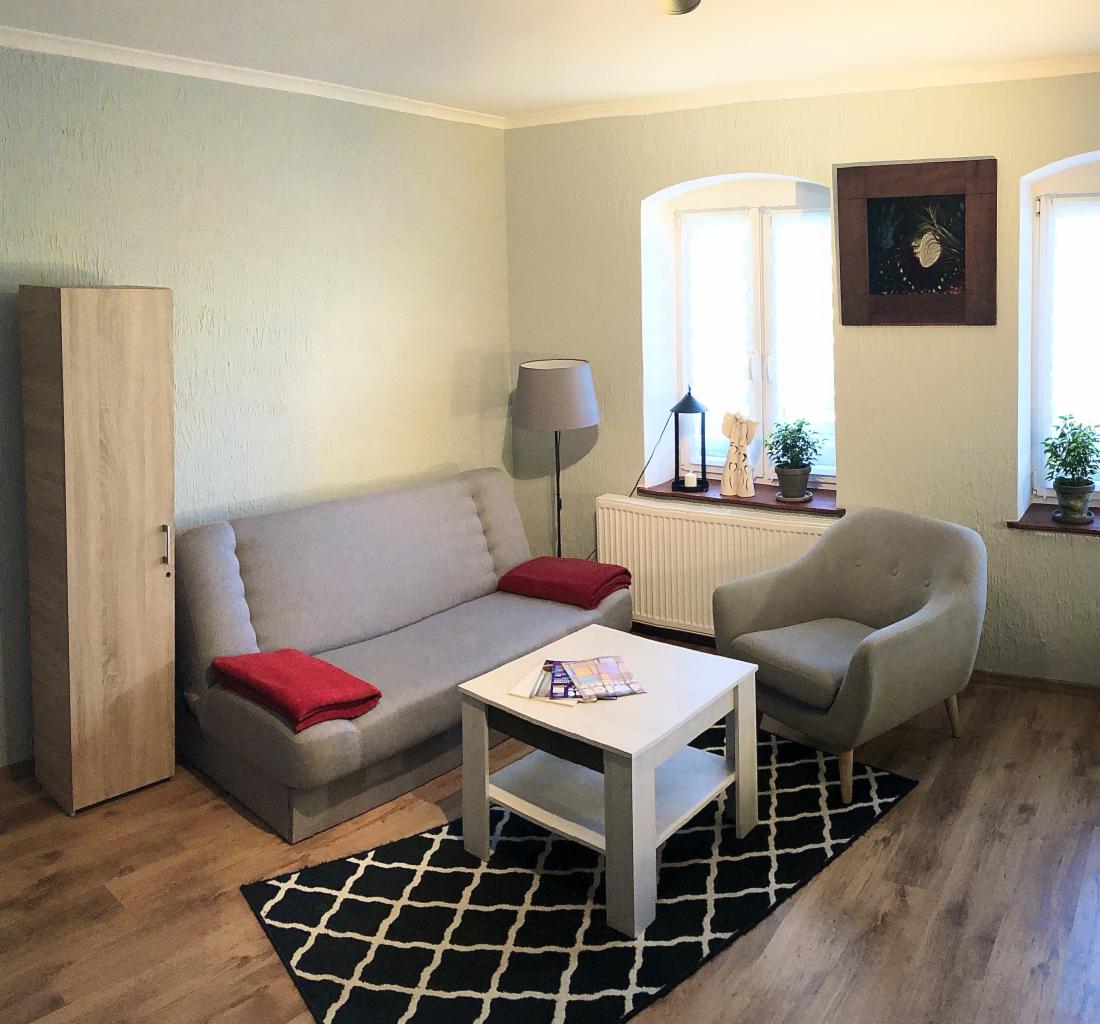 Jasny pokój z szarą sofą z czerwonymi poduszkami, fotelem, białym stolikiem kawowym i czarno-białym dywanem w romby.