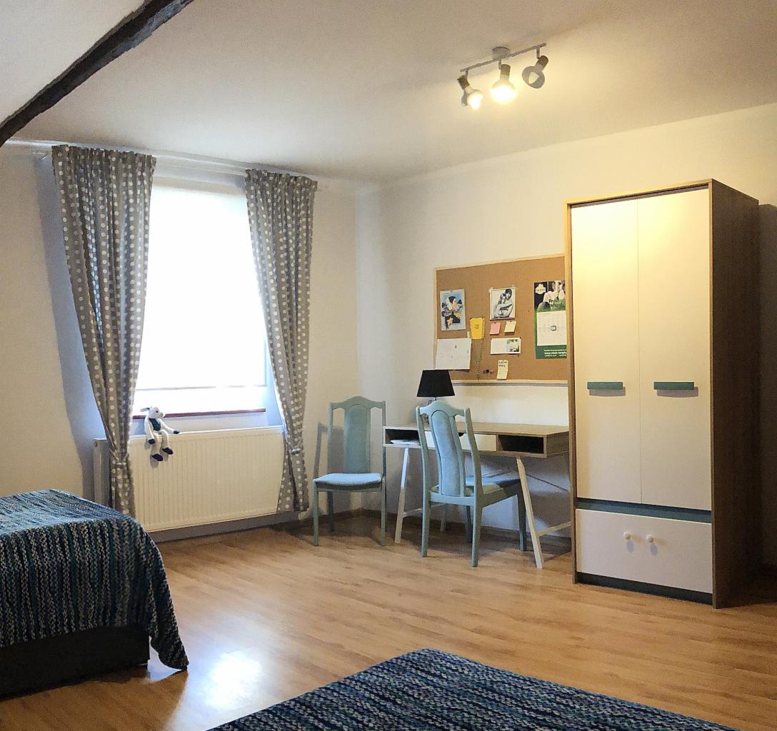 Pokój z dwoma łóżkami, biurkiem, tablicą korkową i szafą, utrzymany w jasnej kolorystyce, z drewnianą podłogą i widocznym fragmentem belki stropowej.