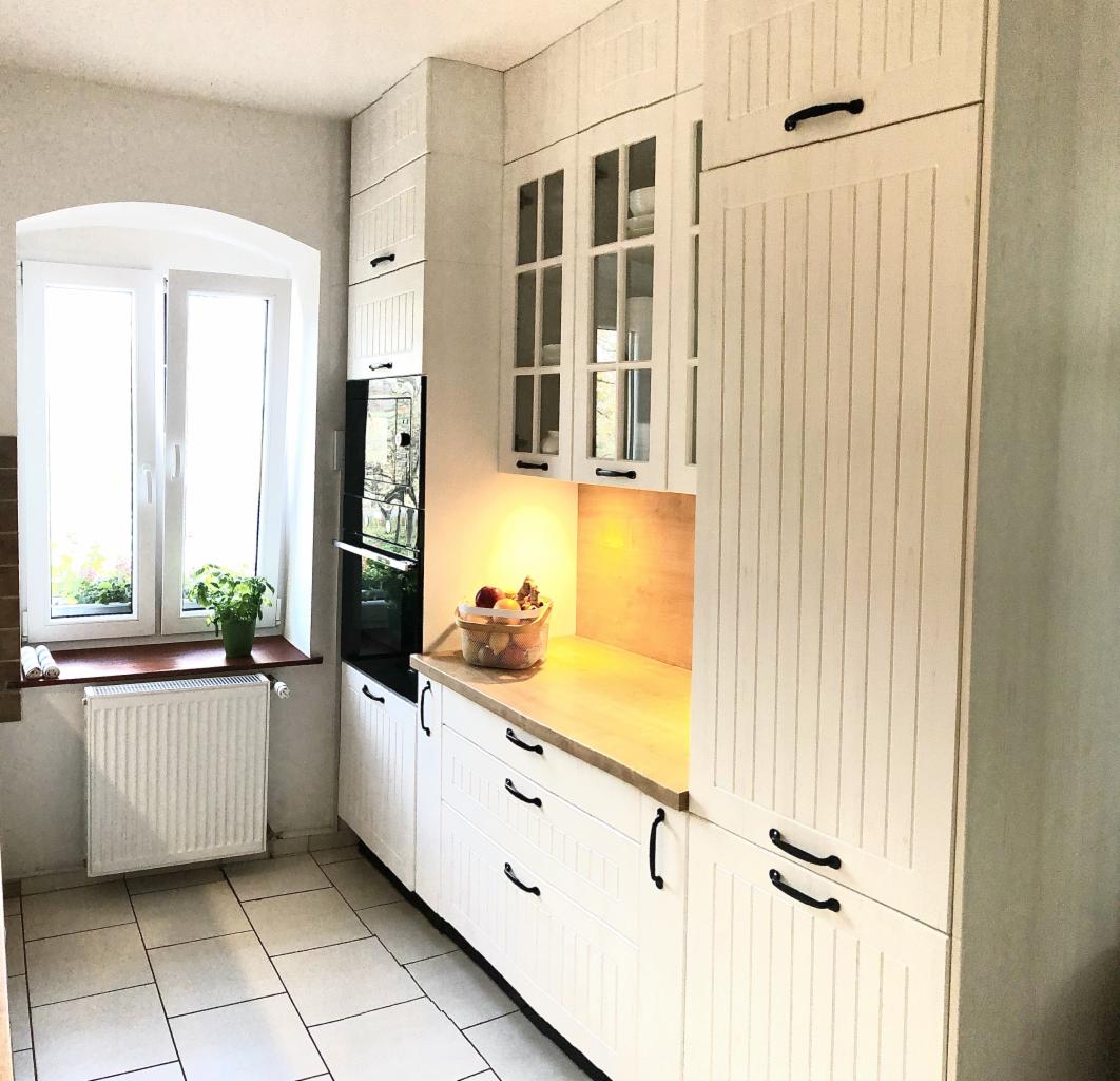 Jasna kuchnia w stylu rustykalnym: białe szafki z frezowanymi frontami i czarnymi uchwytami, drewniany blat, okno z rośliną w doniczce, płytki podłogowe.