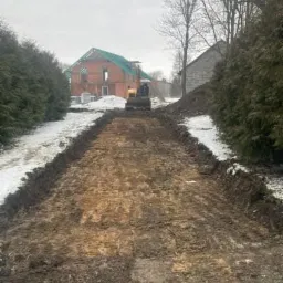 Żółta koparka w tle przygotowuje teren pod budowę drogi na działce z częściowo wybudowanym domem z czerwonej cegły, widoczne ślady śniegu.