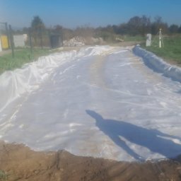 Wilk-trans Maciej Wilk - Przygotowanie podłoża pod drogę: biała geowłóknina rozłożona na ziemi, widoczny cień osoby, w tle budynek i niwelator optyczny.