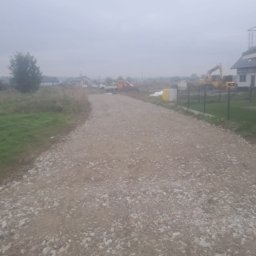 Wilk-trans Maciej Wilk - Szutrowa droga w trakcie budowy na obrzeżach wsi w pochmurny dzień, z widocznymi maszynami budowlanymi w oddali.