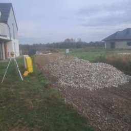 Wilk-trans Maciej Wilk - Kopia kamienia i żwiru przygotowana pod przyszłą drogę na działce budowlanej, widoczny niwelator laserowy i dwa domy w tle.