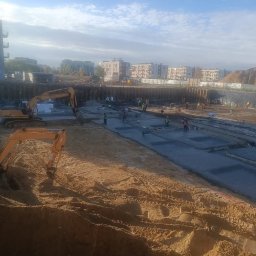 Wilk-trans Maciej Wilk - Dwa żółte koparki na placu budowy z widocznymi fundamentami i pracownikami w oddali, w tle budynki mieszkalne pod błękitnym niebem.