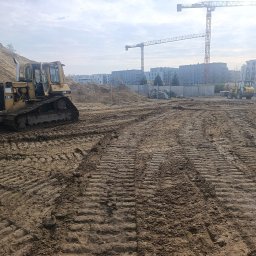 Wilk-trans Maciej Wilk - Żółty spychacz CAT D4H na placu budowy z hałdą piasku w tle, ślady gąsienic na ziemi. W oddali budynki mieszkalne i żurawie budowlane.