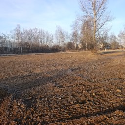 Wilk-trans Maciej Wilk - Przygotowany teren pod budowę z widocznymi śladami maszyn, drzewami w tle i błękitnym niebem. Lekkie oświetlenie słoneczne podkreśla strukturę ziemi.