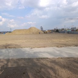 Wilk-trans Maciej Wilk - Rozległy teren budowy z koparką i stertą piasku pod pochmurnym niebem. W tle widoczne budynki i żurawie.