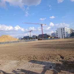 Wilk-trans Maciej Wilk - Teren budowy z hałdą piasku, ogrodzony szarą siatką. W tle bloki mieszkalne i dwa żurawie budowlane na tle błękitnego nieba z chmurami.