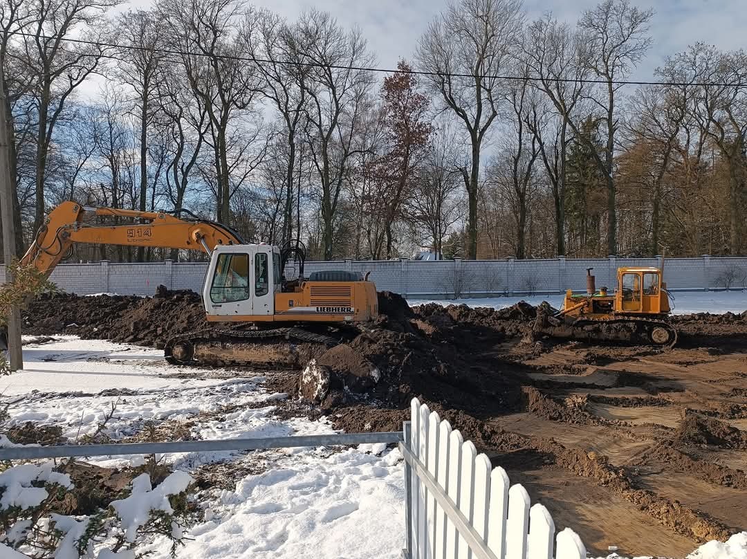 Żółta koparka Liebherr i spycharka pracują na placu budowy w zimowej scenerii, widoczne ślady gąsienic na ziemi, w tle biały mur i drzewa bez liści.