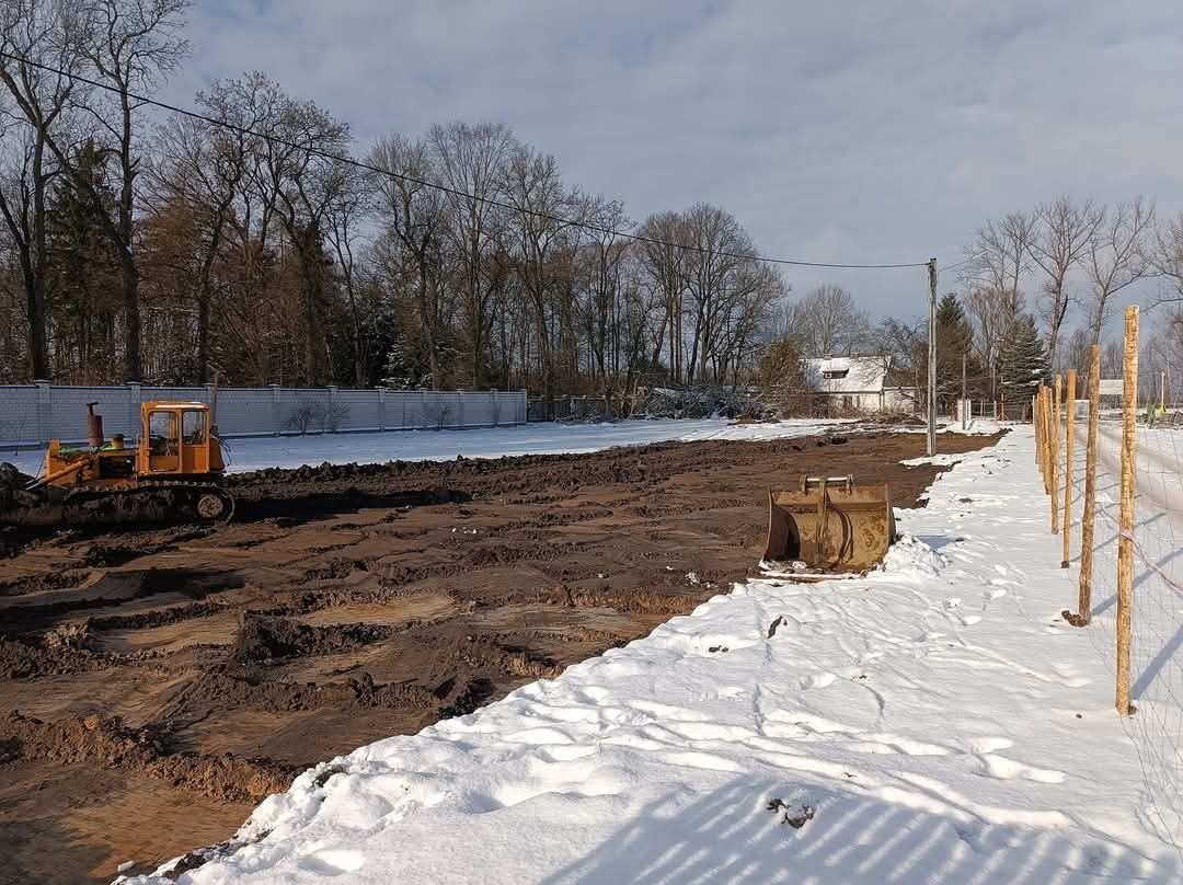 Żółty spychacz gąsienicowy wyrównuje błotnisty teren przylegający do odśnieżonego obszaru, z widocznym wykopem i fragmentem ogrodzenia w tle.